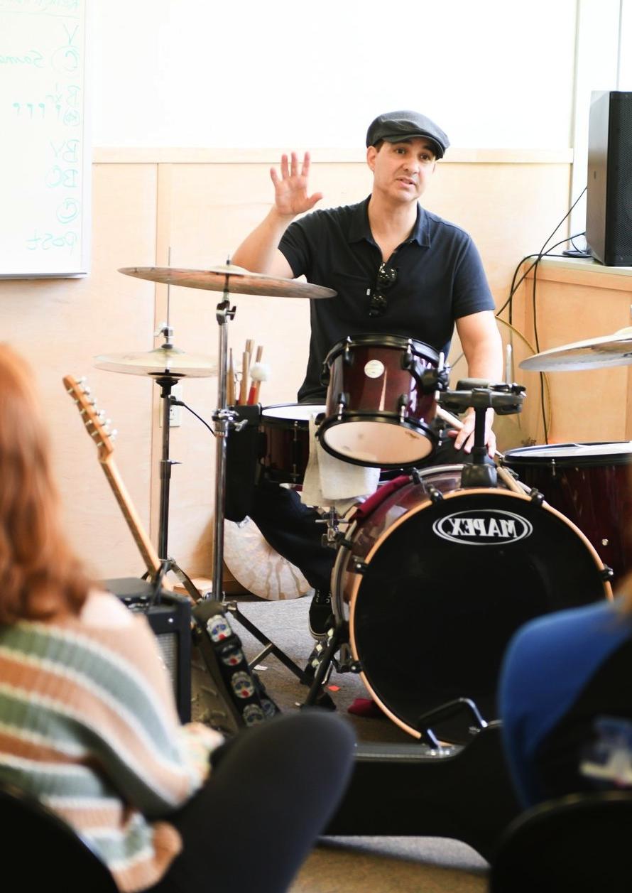 一位身穿黑色衬衫、头戴帽子的音乐教师正坐在架子鼓旁, 在向学生解释的过程中举手. 背景似乎是一个非正式的音乐课，背景是一块白板, 提升教育氛围.